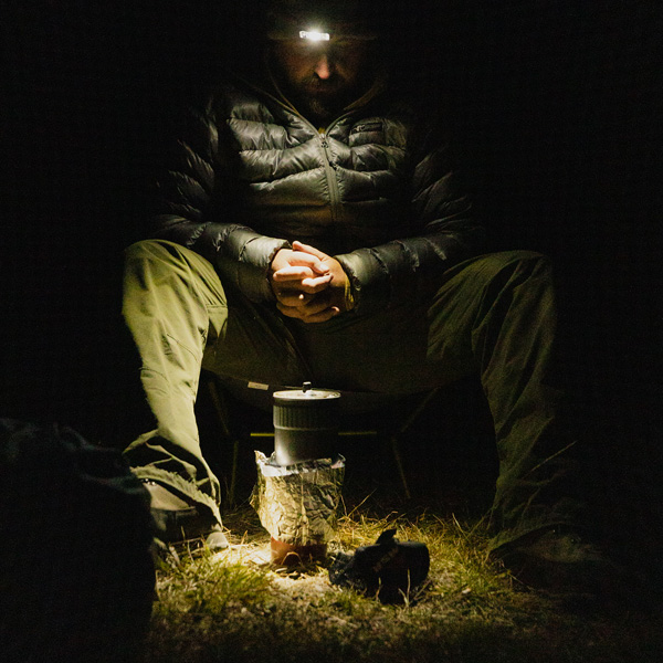Man in the dark with headlamp warming over a small stove 